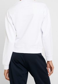 Felpa corta bianca con maniche lunghe, realizzata in un materiale soffice. Presenta un collo tondo e un orlo a coste, abbinata a pantaloni scuri.