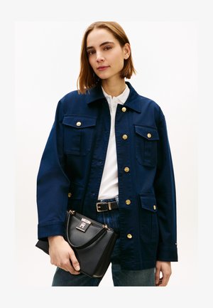 Donna che indossa una giacca blu navy con bottoni dorati, camicia bianca con colletto, jeans blu, che tiene una borsa nera con chiusura dorata.