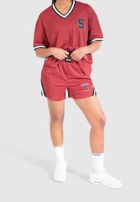 Red athletic shirt with black and white accents, matching red shorts featuring a number and logo, paired with white sneakers and socks.