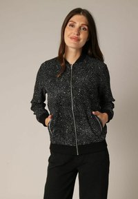 Zwarte bomberjack met een wit gespikkeld patroon, ribgebreide mouwen en zoom, volledige frontale rits en zijzakken. Glad textuur van de stof.