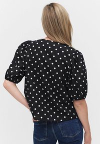 Svart blus med prickar och puffärmar, tillverkad av lätt material. Har vita prickar och en rundad fåll. Bärs med denimjeans.