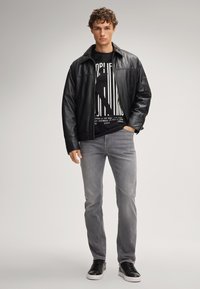 Zwarte leren jasje over een zwart grafisch T-shirt met witte tekst, gecombineerd met lichtgrijze jeans en zwarte sneakers. Eenvoudig ontwerp, casual pasvorm.