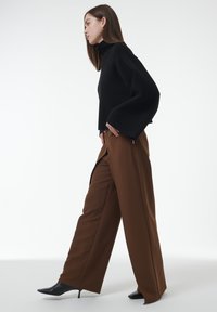 Schwarzer Pullover mit hohem Kragen und weiten Ärmeln, kombiniert mit hoch taillierten, braunen weit geschnittenen Hosen und schwarzen spitzen Pumps. Minimalistisches Design.