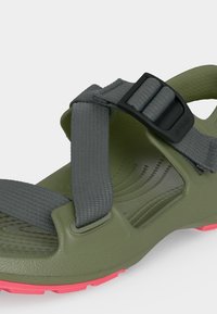 Σανδάλι ελιάς με υφασμάτινους γκρι ιμάντες που μπορούν να προσαρμοστούν, μαύρη πλαστική πόρπη και κόκκινο πέλμα στη κάτω πλευρά.