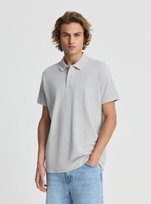 Giovane uomo con capelli ricci che indossa una polo a maniche corte grigio chiaro e jeans azzurri, in piedi con una mano in tasca.