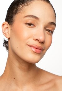 Close-up of a face with smooth, light skin; brown eyes; natural makeup; light brown lips; and short, curly hair, against a white background.