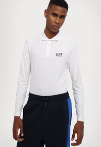 Hvit langermet poloshirt med krage og tre knapper, med svart EA7-logo på brystet, kombinert med mørke joggebukser.