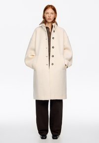 Mujer pelirroja con un abrigo de lana crema de gran tamaño, suéter marrón, pantalones anchos negros y zapatos negros, de pie contra un fondo blanco.