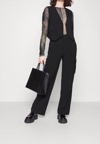 Schwarze ärmellose Weste über einem transparenten Langarmoberteil, kombiniert mit weiten schwarzen Hosen und klobigen schwarzen Schuhen, hält eine kleine schwarze Tasche.