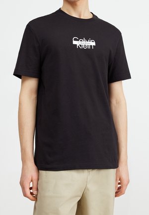 Personne portant un t-shirt noir à manches courtes avec le logo blanc Calvin Klein, associé à un pantalon beige, sur un fond blanc uni.