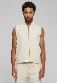 Beige ermeløs vest med glidelås foran, to frontlommer og frynsete kanter. Har "KANI"-logo i hvit søm på skulderen.