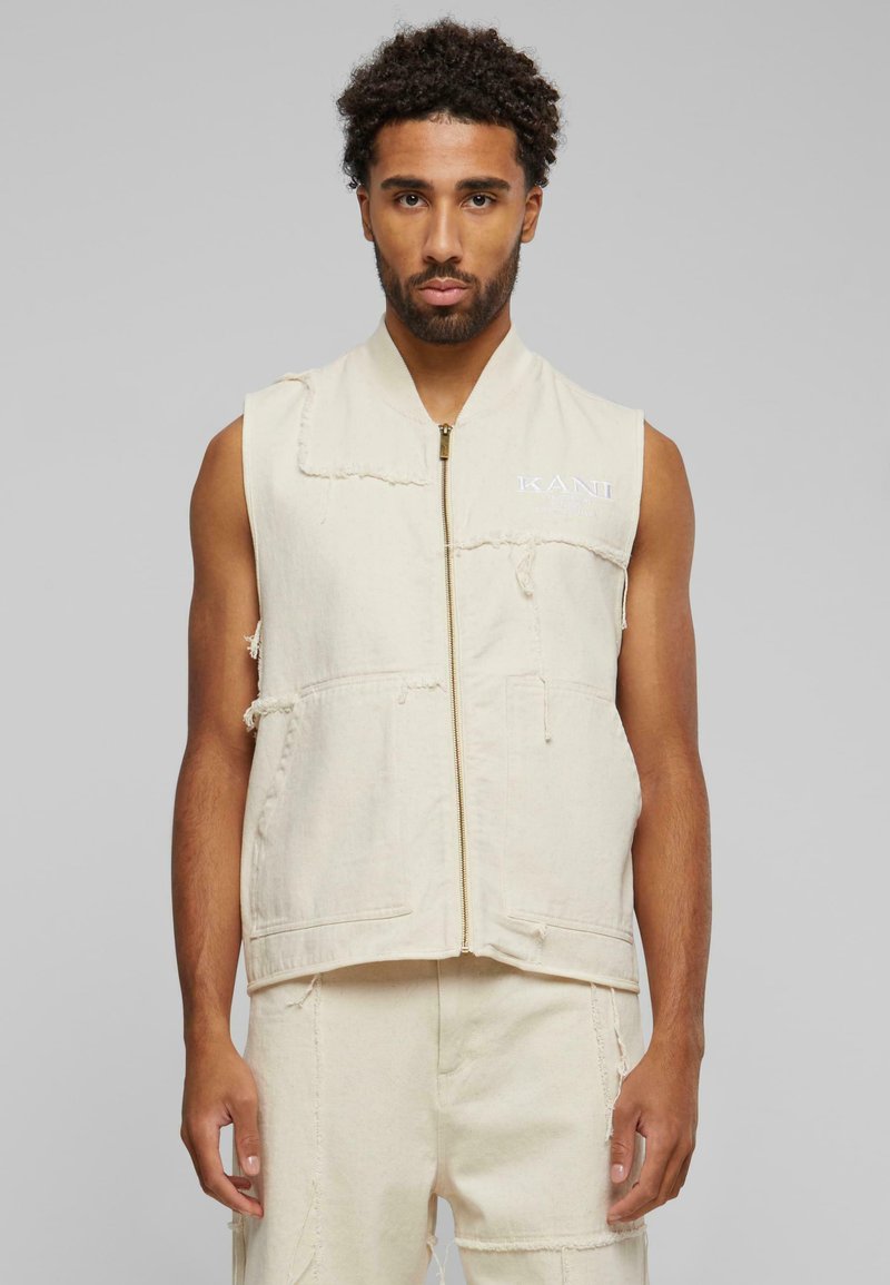 Beige ermeløs vest med glidelås foran, to frontlommer og frynsete kanter. Har "KANI"-logo i hvit søm på skulderen.