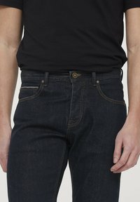 Jean en denim bleu foncé slim, avec des surpiqûres orange, cinq poches et une fermeture à bouton à l'avant. Le matériau présente la texture classique du denim.