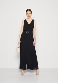 Jumpsuit negro con plisados, escote en V profundo, cintura ajustada y pantalones anchos. Cuenta con un cinturón con una hebilla circular y una textura transparente.