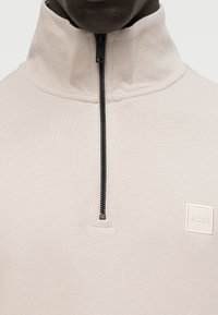 Πουλόβερ με φερμουάρ μέχρι το στήθος σε ανοιχτό μπεζ πλεκτό ύφασμα με υφή και μαύρο φερμουάρ. Διαθέτει μικρό κεντημένο λογότυπο στο στήθος.