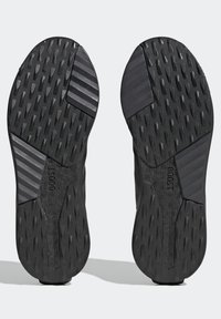 Černé podrážky atletických bot s texturovaným vzorem, kombinující hladkou a vroubkovanou pryž, označené nápisy "ADIWEAR" a "BOOST".