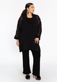 Tunique noire avec un col en cowl, manches longues transparentes et ourlet superposé. Associée à un pantalon noir assorti et des sandales à talons en fourrure noires.