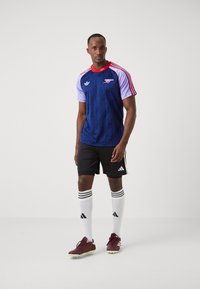 Maglia da calcio Adidas in blu navy e viola con accenti rossi, caratterizzata da un design a pattern, abbinata a pantaloni corti neri, calzettoni bianchi e scarpe da calcio bordeaux.