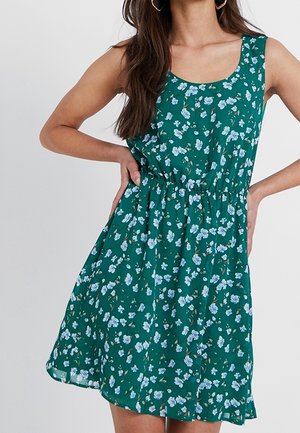 Kvinde iført ærmeløs grøn kjole med lyst blå blomsterprint, markeret talje og A-formet skørt, hænderne hvilende på hofterne.