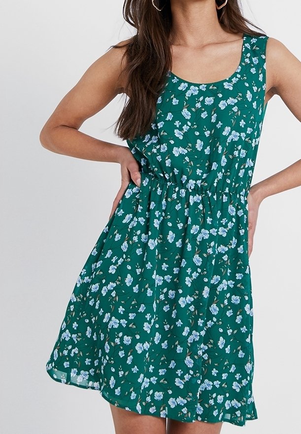 Femme portant une robe verte sans manches avec un motif floral bleu clair, taille cintrée et jupe évasée, mains posées sur les hanches.