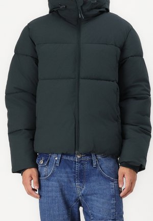 Czarna kurtka puchowa z kapturem i przednim zamkiem błyskawicznym, noszona na niebieskich dżinsach przez stojącą osobę na jednolitym tle.