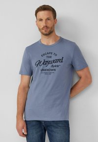 Man met kort haar en baard die een blauw T-shirt draagt met de tekst "Escape to the Wayward Spirit Unknown", staande tegen een effen muur.