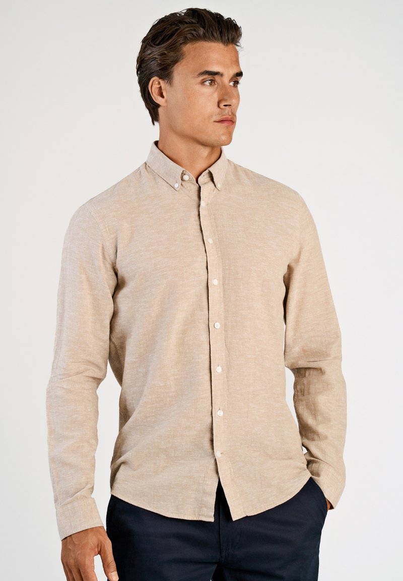 Beige overhemd met knopen en lange mouwen, gemaakt van een gestructureerde stof; voorzien van een kraag en knopen aan de voorkant. Gecombineerd met donkere broek.