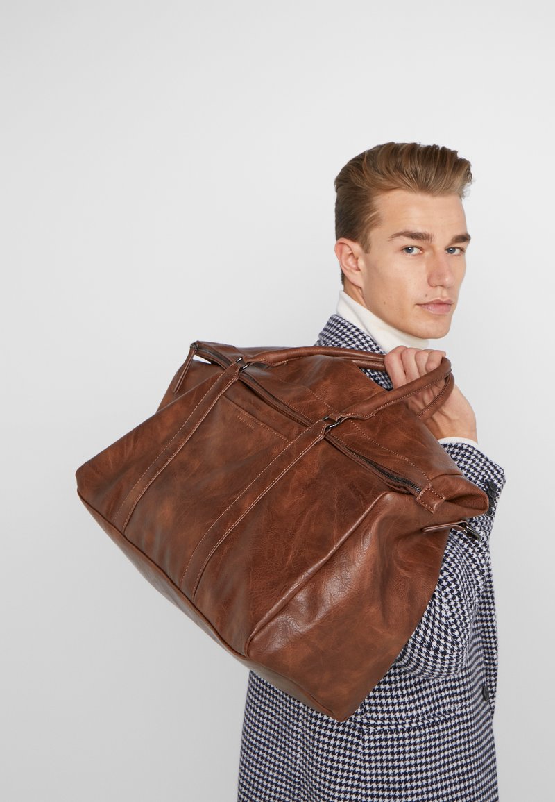 Braune Leder-Duffel-Tasche mit zwei Reißverschlussfächern und strukturiertem Finish, ausgestattet mit zwei Griffen und sichtbaren Nähtelementen.