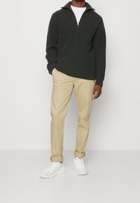 Dunkelgrüner, gerippter Strickpullover mit einem Reißverschlusskragen, kombiniert mit beigen Hosen und weißen Sneakers. Der Stoff hat eine strukturierte Optik.