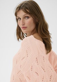 Femme avec des cheveux bruns ondulés portant des boucles d'oreilles en or et un pull en tricot rose avec un motif en vagues, regardant par-dessus son épaule.