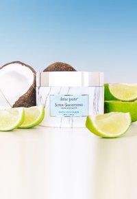 Creamy body butter in a white container with marble pattern, blue label, and silver lid. Fresh coconut and lime slices surround the product.