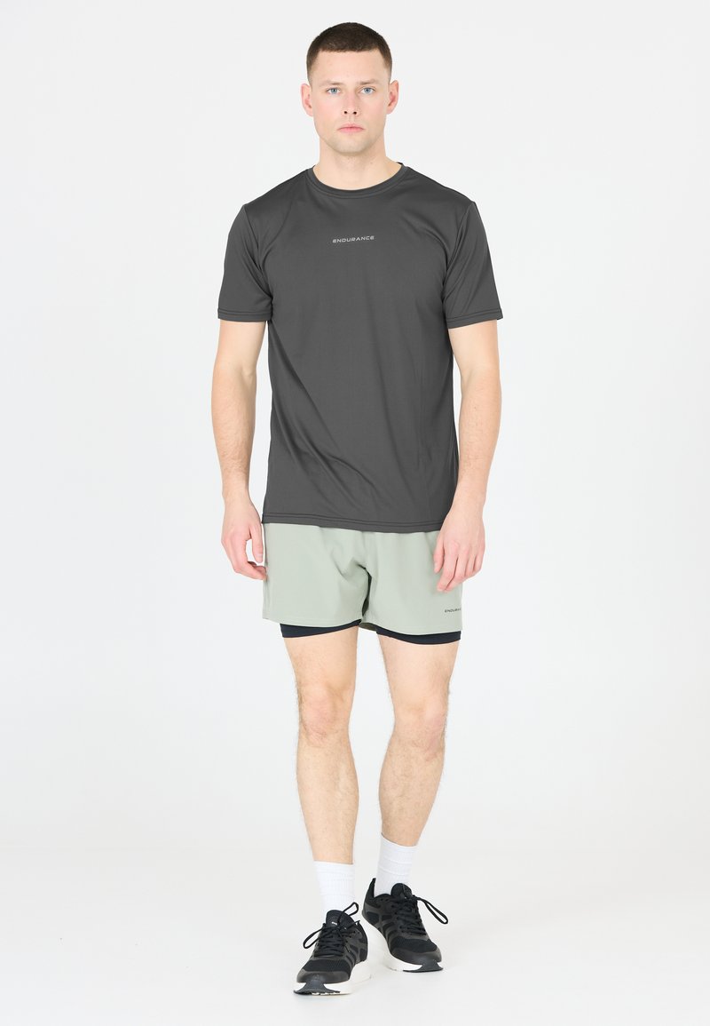 Mörkgrå kortärmad sporttröja med en subtil logotyp, kombinerad med ljusgröna shorts. Modellen har på sig svarta sneakers och vita strumpor.