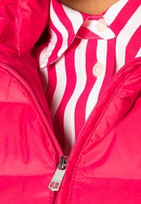 Doudoune rose vif avec une texture brillante et une fermeture éclair argentée, superposée à une chemise à col rayée blanche et rose.