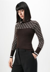 Mujer con pelo lacio y negro, vestida con un suéter marrón ajustado con un cuello y mangas blancas estampadas, pantalones negros, con una mano en el bolsillo.