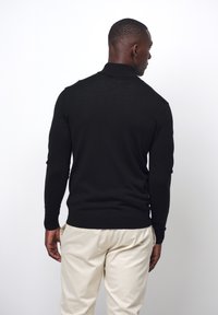 Schwarzer langärmeliger Pullover mit taillierter Passform und Kragen, aus strukturiertem Stoff, getragen mit beigen Hosen, von hinten betrachtet.