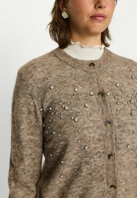 Bruine wollen cardigan met een getextureerd oppervlak en verspreide metallic versieringen. Knopen als sluiting en een ronde halslijn.