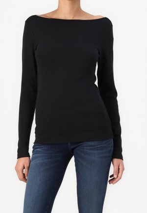 Kobieta w dopasowanym czarnym topie z długim rękawem, szerokim dekoltem w łódkę i niebieskich jeansach na jednolitym białym tle.