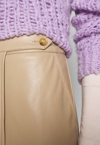 Beige nahkahousut, joissa on sileä pinta, sivusauma ja pyöreä keltainen nappi, yhdistettynä violettiin neulepuseroon.