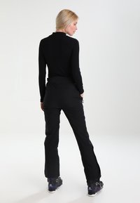 Black long-sleeve top with a high neckline, paired with black pants featuring pockets and zippers at the ankles, worn with dark ski boots.