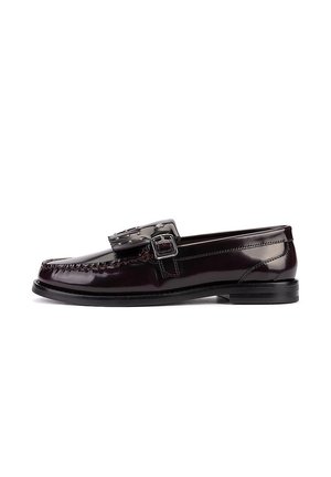 Mocasín de cuero marrón oscuro brillante con suela negra, borla decorativa y correa lateral con hebilla, visto desde el lado exterior.