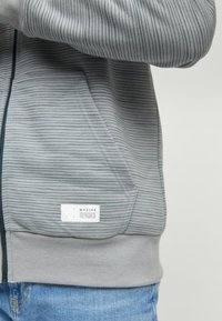 Szara bluza z zamkiem błyskawicznym w cienkie poziome paski, z przednią kieszenią kangurką, biała metka, noszona na niebieskich dżinsach, częściowy widok osoby z uniesioną ręką.