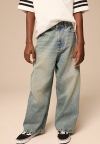 Hellblaue, weite Jeans aus Denim mit einem ausgewaschenen Finish, lockerer Passform und geradem Schnitt, kombiniert mit schwarzen Sneakers.