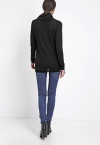 Schwarzes Langarmshirt mit Wasserfall-Ausschnitt, aus einem strukturierten Strickmaterial gefertigt. Kombiniert mit schmal geschnittenen blauen Jeans und schwarzen Absatzstiefeletten.
