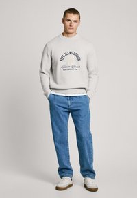 Vaalea harmaa neulepaita, jossa on painettu logo, yhdistetty sinisiin corduroy-housuihin. Mallilla on päällään valkoiset tennarit, joissa on ruskeita yksityiskohtia.