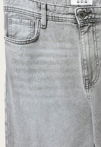 Jeans in denim grigio chiaro che mostrano la tasca frontale, il passante per cintura, il bottone con il marchio e l'etichetta della taglia che indica ITA 46, USA 31, UE 42.