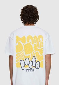 Fehér póló, amelynek hátulján nagy sárga tésztaformák grafika található, és szürke betűkkel a "tészta" felirat, alatta pedig elérhetőségek.