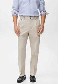 Beige op maat gemaakte broek met voorplooien, zijzakken en een gladde stofstructuur, gecombineerd met zwarte leren schoenen en een lichtblauw gestreept shirt.