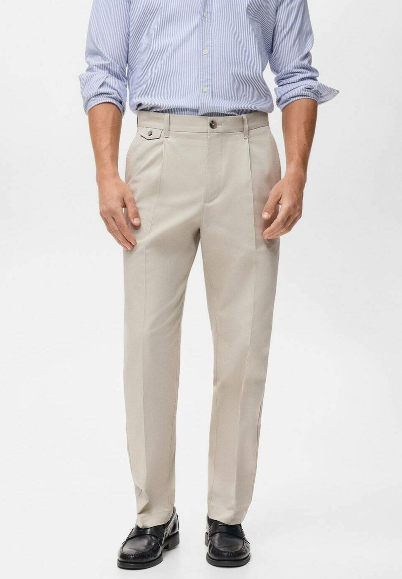 Beige op maat gemaakte broek met voorplooien, zijzakken en een gladde stofstructuur, gecombineerd met zwarte leren schoenen en een lichtblauw gestreept shirt.