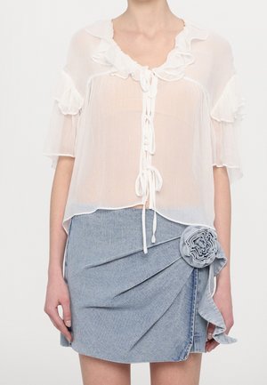 Blouse blanche transparente avec col à volants et liens, associée à une jupe en jean bleu clair ornée d'un grand détail de fleur en tissu.