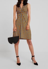 Vestido sin mangas con un patrón geométrico en naranja, negro y blanco, que presenta un diseño de frente retorcido. Combinado con un bolso negro y tacones.